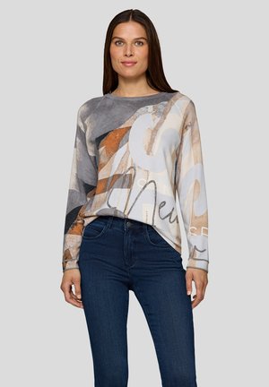Frau mit langen braunen Haaren, die ein langärmliges Oberteil mit abstraktem Muster in Grau-, Beige- und Orangetönen trägt, kombiniert mit dunkelblauen Jeans.