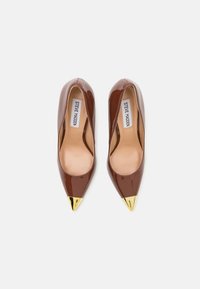 Un par de zapatos de tacón puntiagudos de color marrón brillante con puntas metálicas doradas y plantillas beige, etiquetados como Steve Madden, sobre un fondo blanco.