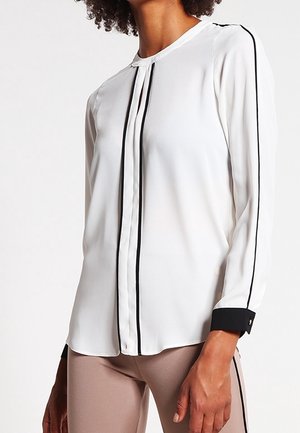 Kobieta ubrana w białą bluzkę z długim rękawem z czarnymi lamówkami na rękawach i z przodu, w połączeniu z beżowymi spodniami z czarnymi paskami po bokach.