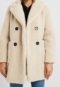 Mujer vestida con un abrigo de teddy texturizado, de color crema y de doble botonadura, con botones negros, sobre una camisa negra y jeans azules.