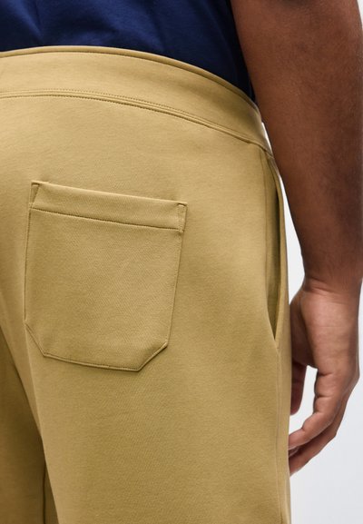 Κοντινό πλάνο ατόμου που φοράει μπεζ παντελόνι με πίσω τσέπη και μπλε σκούρο πουκάμισο, δείχνοντας την πλευρική και την πίσω περιοχή της μέσης.