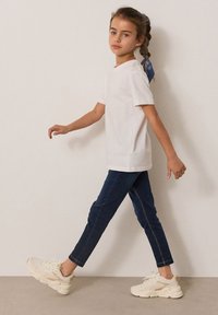 Bijela kratka majica s kratkim rukavima, tamne uske traperice i kremaste tenisice s svijetlim naglascima. Kosa oblikovana plavom maramom.