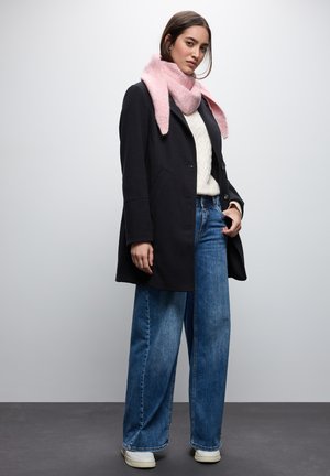 Cappotto di lana nero sopra un maglione bianco a trecce, abbinato a jeans blu a gamba larga e una sciarpa rosa. Sneakers bianche completano l'outfit.