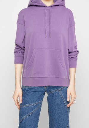 Personne portant un sweat à capuche violet ample avec cordons et poche avant, associé à un jean bleu avec des détails de coutures diagonales.