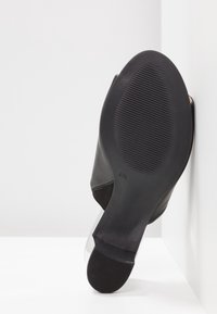 Svart sko med en slät läderöverdel, en genomskinlig klack och en texturerad gummisula med horisontella spår.