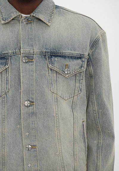 Light blue denim jacket with silver-tone buttons, two chest pockets, and decorative stitching in a cross pattern. Distressed texture detail.