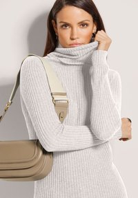 Frau in einem hellgrauen, gerippten Rollkragenpullover, die den Kragen hochhält, mit einer beigen Umhängetasche mit breitem Tragegurt über der Schulter.