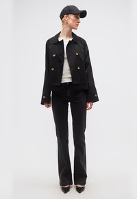 Άτομο με μαύρο καπέλο, μαύρο cropped διπλό σακάκι, άσπρο πουκάμισο, μαύρα τζιν και μαύρα μυτερά παπούτσια, στέκεται μπροστά σε λευκό φόντο.