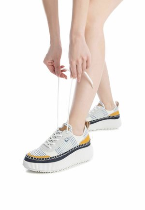 Zapatillas blancas y azules con un patrón, suela blanca gruesa y detalles en amarillo y negro. Persona atándose los cordones.