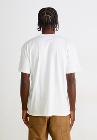 Tricou alb cu guler rotund și mâneci scurte, confecționat dintr-un material neted. Purtați cu pantaloni scurți maro tip cargo. Vedere din spate, fără grafice.