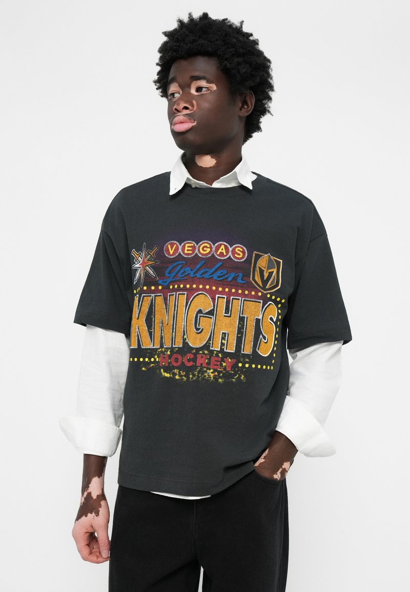 Dunkelgraues kurzärmeliges T-Shirt mit buntem "Vegas Golden Knights" Grafikdruck, der Sterne und Punkte zeigt, kombiniert mit einem weißen Hemd mit Kragen darunter.