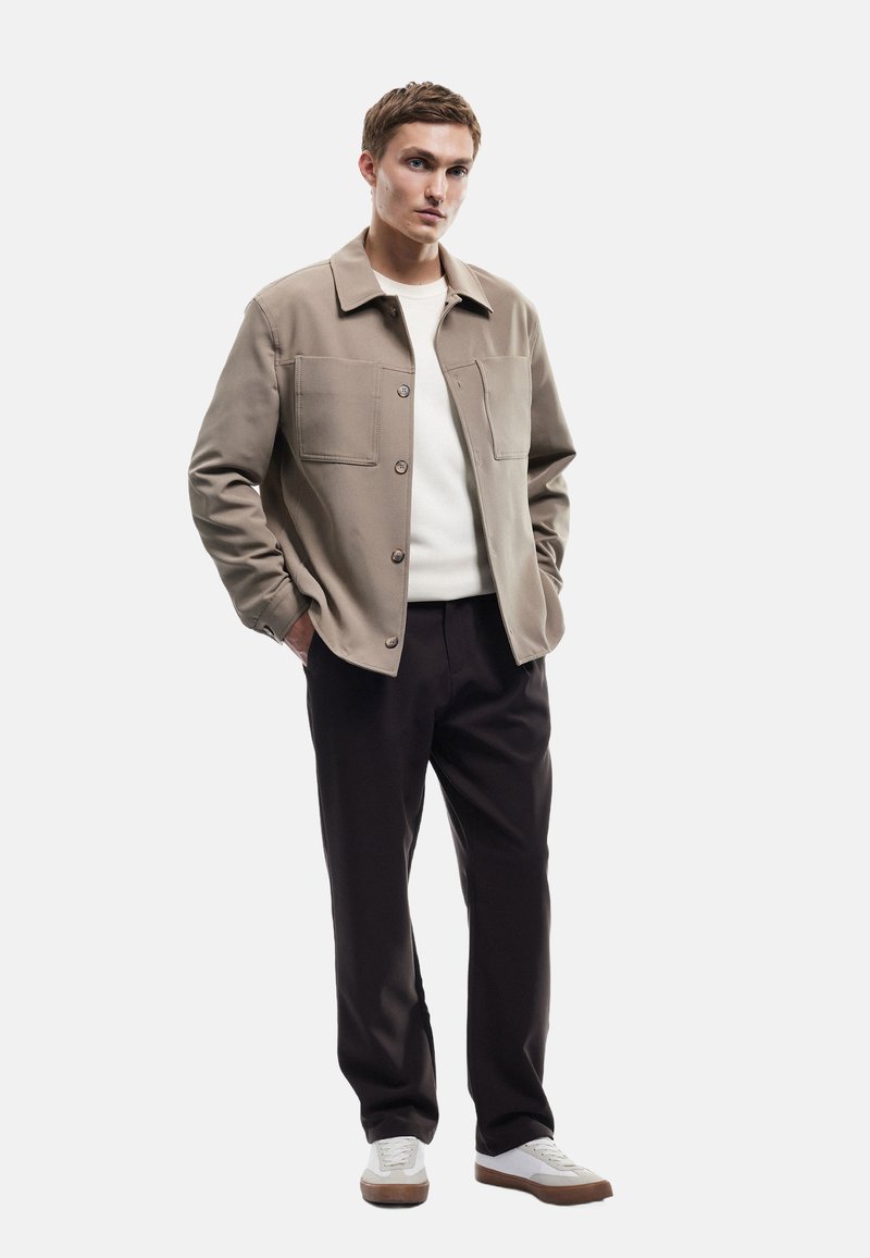 Beige overshirt met twee borstzakken, gecombineerd met een witte trui en donkere broeken. Model draagt witte sneakers met bruine zolen.