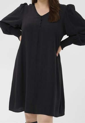 Kobieta w czarnej sukience z długim rękawem z bufkami, ściągaczami na mankietach i trzema małymi guziczkami na piersi, stojąca z rękami na biodrach.