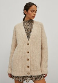 Beige gebreide cardigan met een V-hals, vijf houten knopen en een ribbeltextuur. Draagt over een zwart en beige slangenprintjurk.