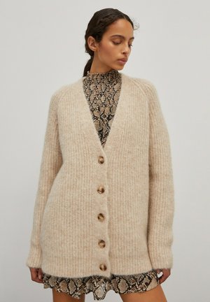 Beige gebreide cardigan met een V-hals, vijf houten knopen en een ribbeltextuur. Draagt over een zwart en beige slangenprintjurk.