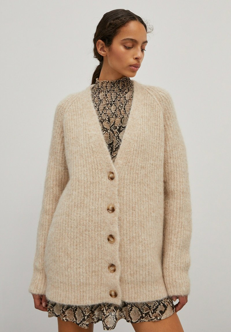 Beige gebreide cardigan met een V-hals, vijf houten knopen en een ribbeltextuur. Draagt over een zwart en beige slangenprintjurk.