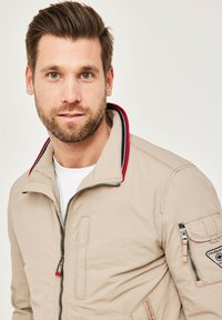 Beige bomber jacket with a red and blue striped collar, zippered pockets, and a textured finish. Features a logo patch on the sleeve.