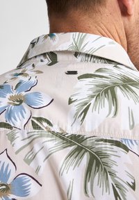 Camisa ligera con estampado floral en beige, que presenta flores azules y hojas verdes. Incluye una sutil etiqueta de marca encima del cuello.