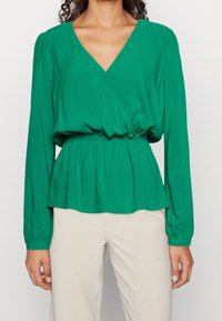 Blusa verde avvolgente con scollo a V, maniche lunghe a palloncino, cintura elastica e una silhouette fluida. Tessuto morbido con una superficie liscia.