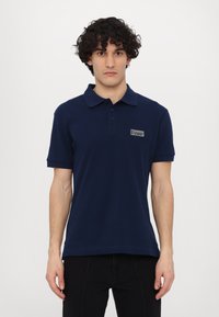 Polo majica temno modre barve iz bombaža, s klasičnim ovratnikom, kratkimi rokavi, tri gumbi na preklopu in našitkom z logotipom na prsih.
