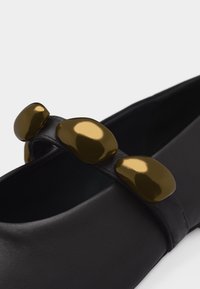 Μαύρο επίπεδο παπούτσι με λουράκι διακοσμημένο με τρεις λαμπερές, ακανόνιστου σχήματος χρυσές διακοσμήσεις κατά μήκος της κορυφής.