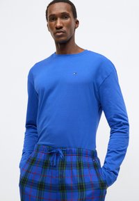 Modrá košeľa s dlhým rukávom a okrúhlym výstrihom, v páre so zeleno-modrými károvanými pyžamovými nohavicami s pásom na sťahovanie. Červené akcenty na páse.