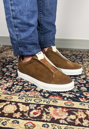 Baskets en daim marron à enfiler avec semelles blanches, portées avec un jean bleu, sur un tapis à motifs floraux multicolores.