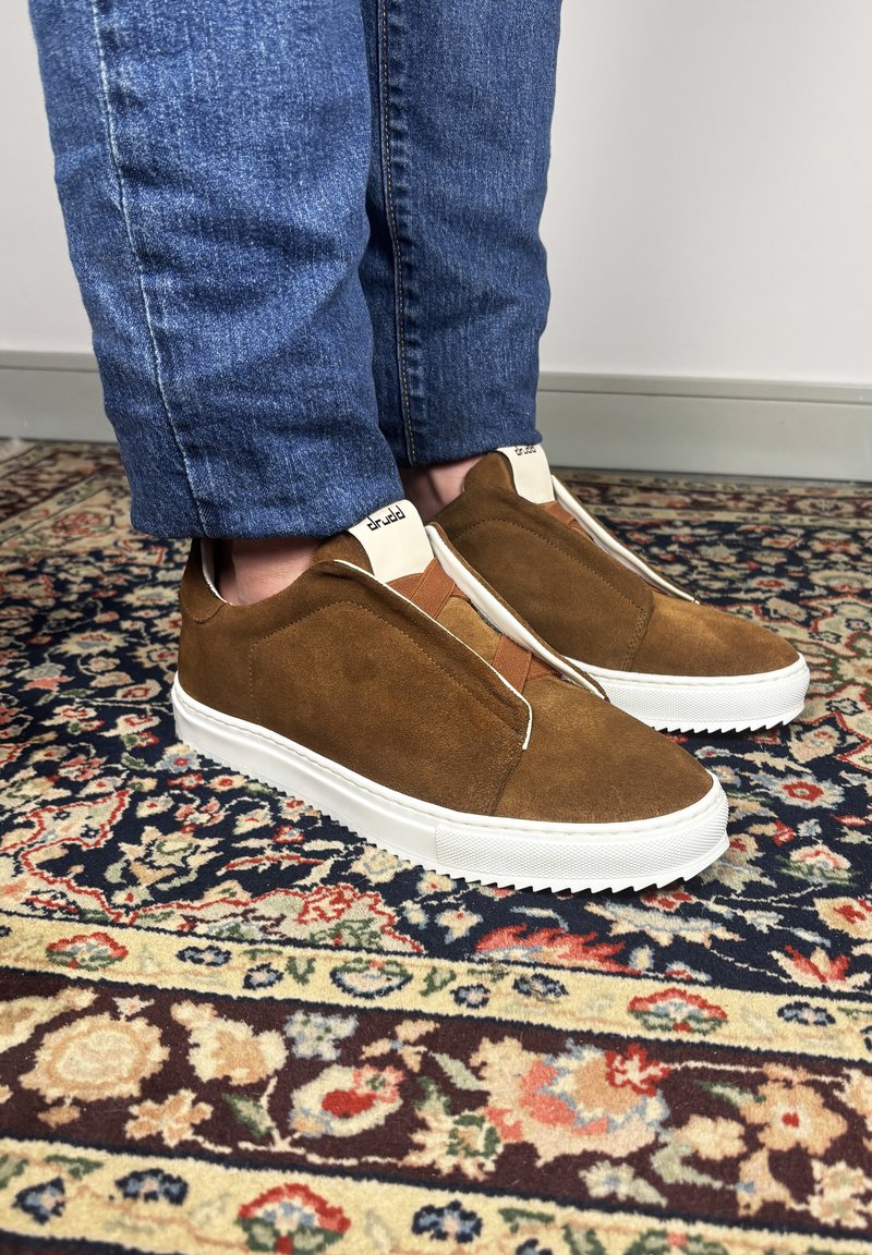 Bruine suède slip-on sneakers met witte zolen, gedragen met een blauwe spijkerbroek, staand op een meerkleurig vloerkleed met bloemmotief.