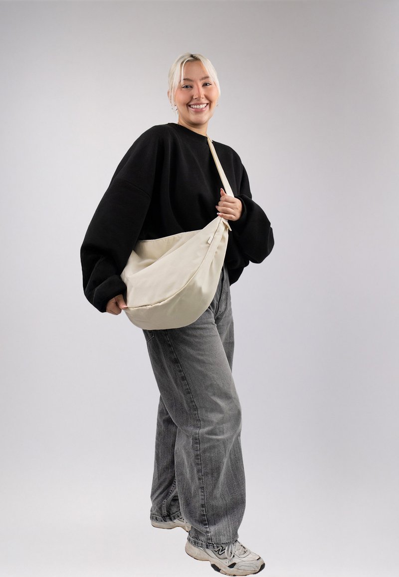 Borsa a tracolla beige in tela; morbida al tatto, dalla forma floscia, con una sola tracolla indossata su una spalla su uno sfondo grigio.