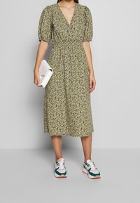 Midi šaty s květinovým potiskem, V výstřihem, nafouknutými rukávy a přiléhavým pasem, ve žluté a námořnické modři, ve spojení s bílými teniskami a bílou kabelkou.