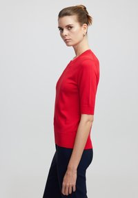 Suéter rojo de manga corta con cuello redondo, textura suave y diseño ajustado. Combinado con pantalones oscuros. Fondo simple.
