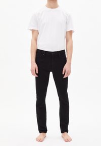 Jean en denim noir avec une coupe slim, présentant une fermeture à boutons, un design à cinq poches et une texture lisse. Associé à un t-shirt blanc uni.