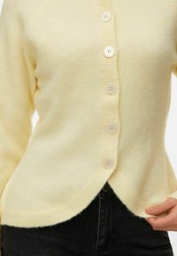 Cardigan jaune doux avec six boutons porté sur un pantalon noir, visible de la taille au milieu de la poitrine, une main tenant l'ourlet.