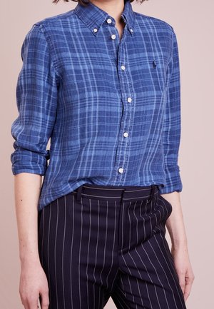 Person, die ein blau kariertes Hemd mit hochgekrempelten Ärmeln und schwarze Nadelstreifenhose vor einfachem Hintergrund trägt.