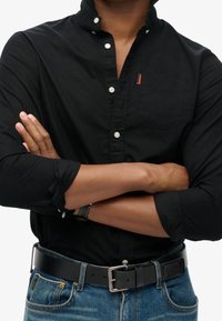 Camicia nera con colletto e taschino sul petto, decorata con un piccolo logo rosso. Indossata con jeans scuri e una cintura nera.