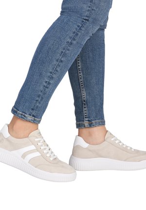 Jambes en jean bleu slim portant des baskets beige et blanches à lacets avec des semelles blanches épaisses, debout sur un fond blanc.