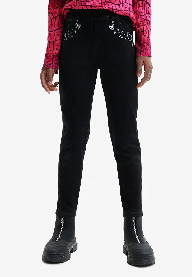 Personne portant un pantalon slim noir avec une broderie blanche de cœur et d’étoile près des poches, des bottes noires épaisses avec fermetures éclair à l’avant et un haut rouge à motifs.