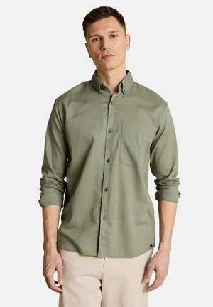 Junger Mann trägt ein hellolivgrünes Hemd mit hochgekrempelten Ärmeln und beige Hose, steht vor einem schlichten weißen Hintergrund.