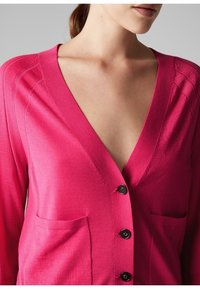 Roze gebreide cardigan met een diepe V-hals, zwarte knopen en twee voorzakken. De stof lijkt glad met een gemiddelde textuur.