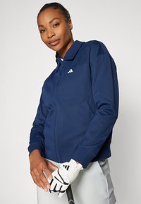 Tamsiai mėlynas zip-up džemperis su apykakle, turintis mažą baltą logotipą ir šonines kišenes, derinamas su baltais golfų pirštinėmis, prieš neutralų foną.