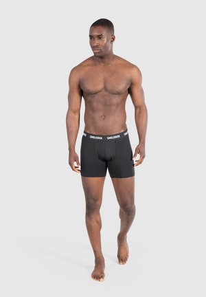 Boxers noirs avec une texture lisse et une taille élastique arborant le logo "SMILODON" en blanc. Modèle debout pieds nus, fond neutre.