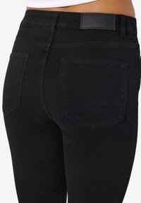 Femme portant un jean noir moulant montrant les poches arrière et la zone de la taille avec un top court blanc.