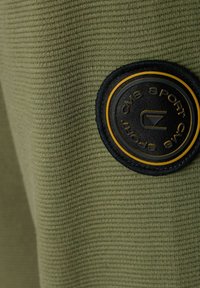 Olive green, teksturowana tkanina z okrągłą czarną łatką z żółtymi akcentami, na której widnieje logo w stylizowanej czcionce.