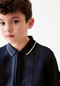 Jonge jongen met krullend donker haar, die een navyblauw gebreid poloshirt draagt met een wit afgezette kraag, tegen een effen witte achtergrond.