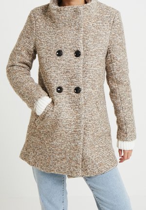 Femme portant un manteau en laine beige texturé à double boutonnage avec des boutons noirs, des poignets en tricot blanc, et un jean bleu clair, main dans la poche du manteau.