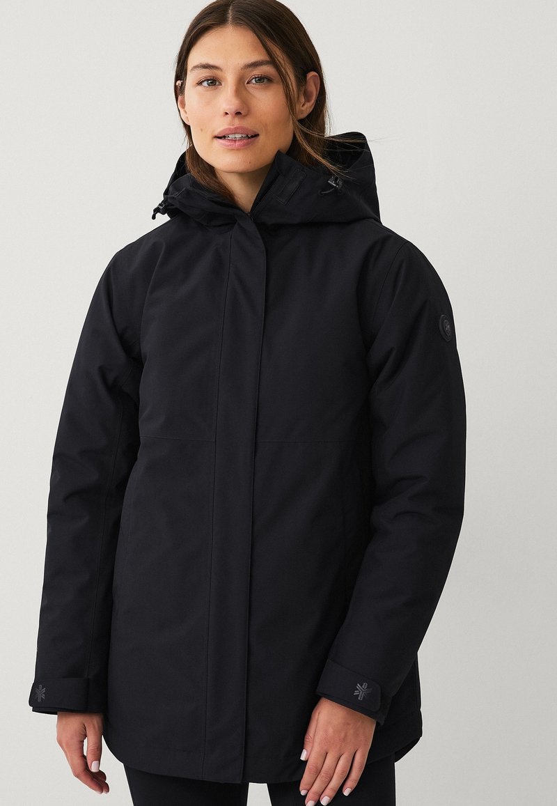 Vrouw in een zwart gewatteerd winterjack met capuchon, een rits en klittenbandsluitsingen, staande tegen een effen lichte achtergrond.