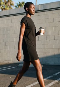 Junge Frau in einem schwarzen Kleid und Stiefeln geht draußen spazieren und hält einen To-Go-Kaffeebecher vor einem grauen Wandhintergrund.