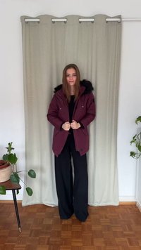 Parka bordeaux avec capuche doublée de fourrure, fermetures éclair et boutons-pression, poches latérales ; portée avec un pantalon noir, sur un fond de rideau neutre.