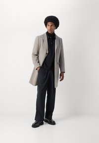 Abrigo de lana gris con polo negro y pantalones de rayas. Zapatos negros. Modelo de pie con las manos en los bolsillos frente a un fondo sencillo.