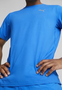 Modrý sportovní tričko vyrobené z hladké, lehké látky. Má kulatý výstřih a malou zlatou značku Puma na hrudi.